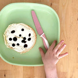 A child's hand reaches for a pink stainless steel child safe knife. A round slice of bread sits on the green plate with white spread.