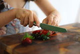 A child cuts strawberries on a wooden cutting board with a sea green stainless steel child safe kitchen knife.